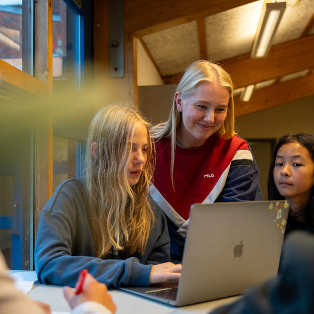 Elever og lærer snakker sammen om en oppgave på macbook.