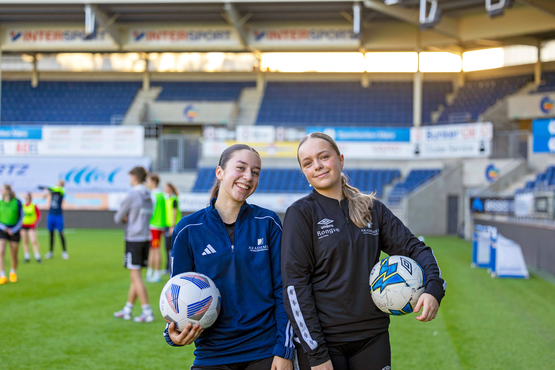 To jenter står på fotballbanen med hver sin fotball under armen.