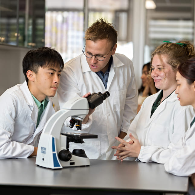 Tre elever og en lærer har på labfrakker og ser inn i et mikroskop i undervisningen.