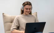 Jente som smiler mens hun jobber med fag på Mac med headset. Foto.