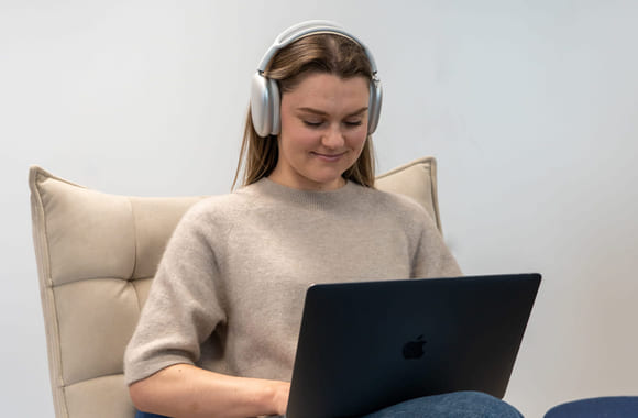 Jente som smiler mens hun jobber med fag på Mac med headset. Foto.