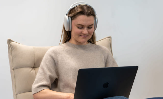 Jente som smiler mens hun jobber med fag på Mac med headset. Foto.