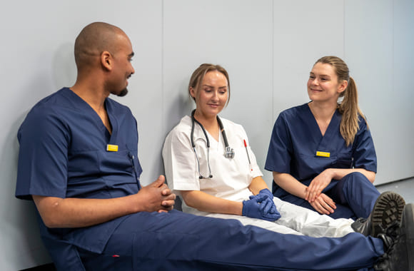 Tre helsefagarbeider som sitter og snakket sammen og smiler. Foto.