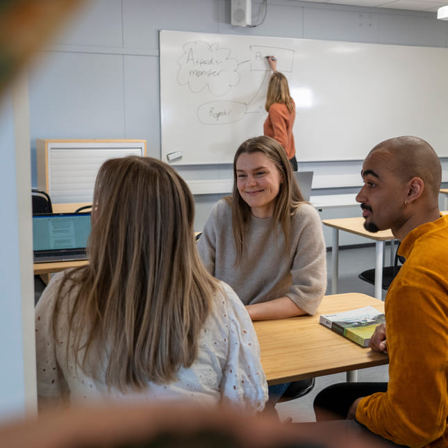 Tre elever sitter i et klasserom med lærer i bakgrunn