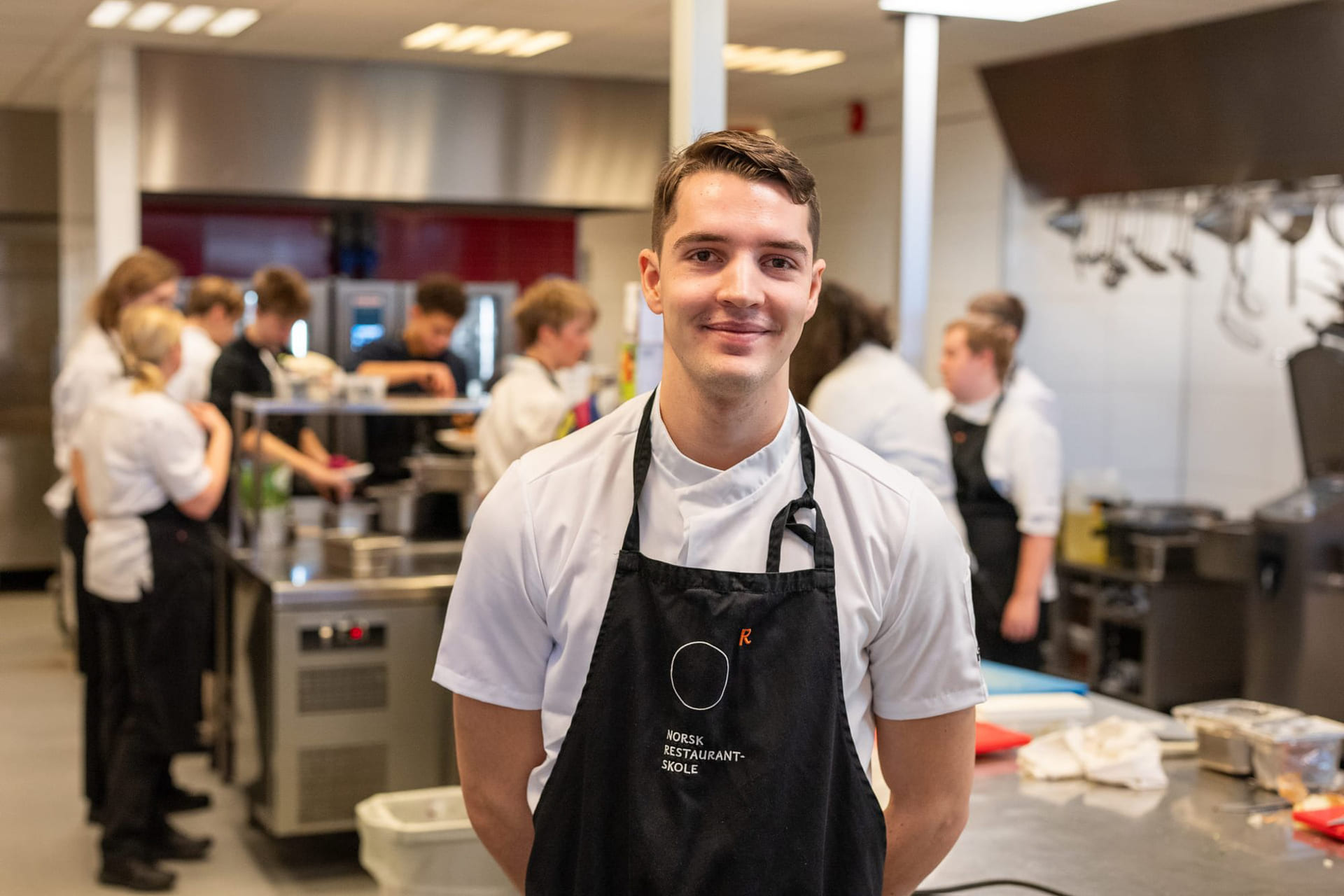 Elev på Akademiet norsk Restaurantskole smiler til kamera med klassekamerater i bakgrunnen
