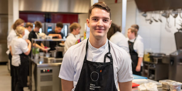 Elev på Akademiet norsk Restaurantskole smiler til kamera med klassekamerater i bakgrunnen