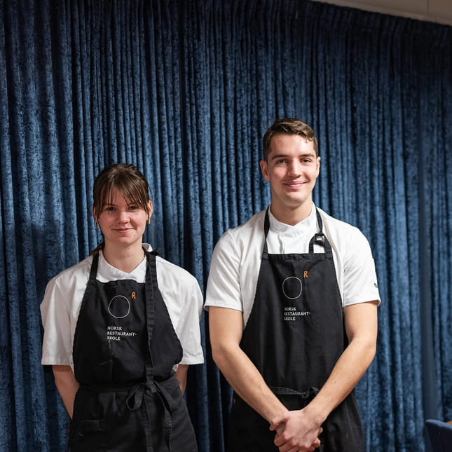 To elever på akademiet norsk restaurantskole poserer foran et dekket bord.